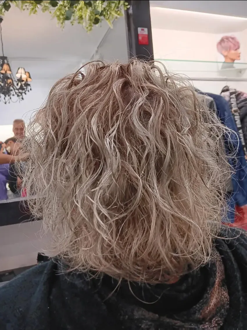 Espalda de una persona con cabello rizado y gris en una peluquería, con espejos y percheros al fondo.