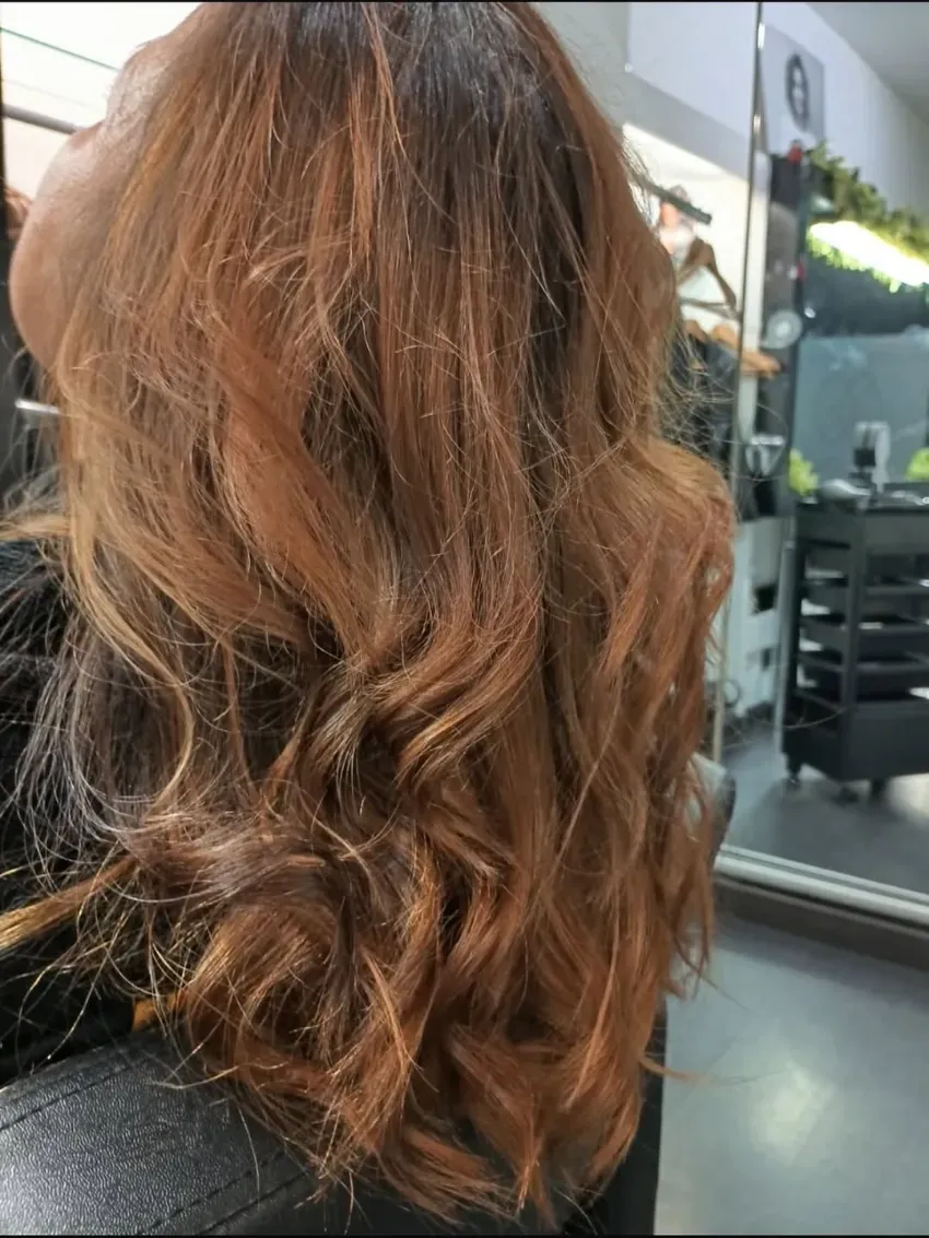 Cabello largo, ondulado y castaño rojizo, peinado con rizos sueltos, visto desde atrás en una silla de peluquería.