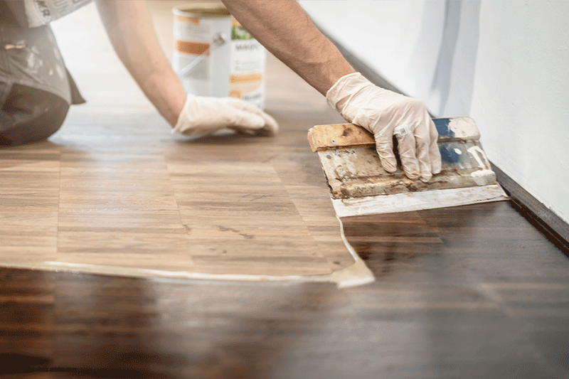 Eine Person mit Handschuhen trägt mit einer Kelle Dichtmittel auf einen Holzboden auf.
