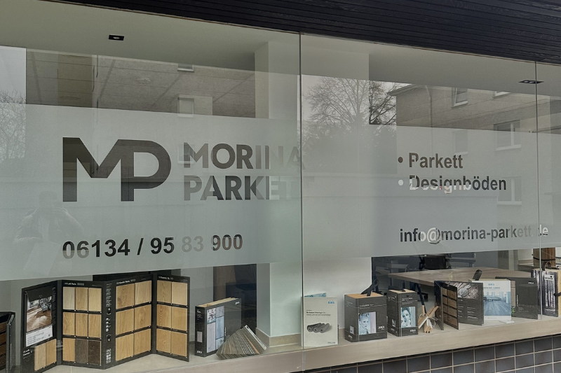 Schaufenster von Morina Parkett mit Logo, Telefonnummer und ausgestellten Bodenbelagsmustern.