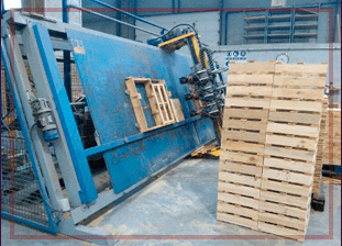 Máquina industrial azul ensamblando palés de madera. Pila de palés junto a la máquina.