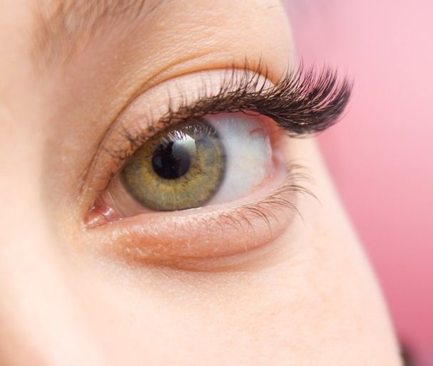 Primer plano de un ojo con pestañas largas y oscuras sobre un fondo rosa. El iris es verde.