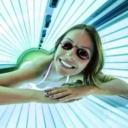 Una mujer que usa gafas bronceadoras sonríe en una cama bronceadora, rodeada de luz azul.