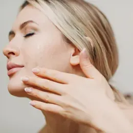 Mujer con cabello rubio, ojos cerrados, aplicándose un producto de cuidado de la piel en la mejilla con la mano.