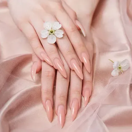 Primer plano de unas manos con uñas largas, almendradas y de color nude, adornadas con un anillo de flor blanca.