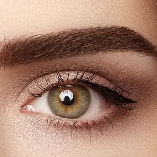Primer plano de un ojo humano con iris color avellana verde-marrón, cejas definidas y una sutil sombra de ojos neutra.