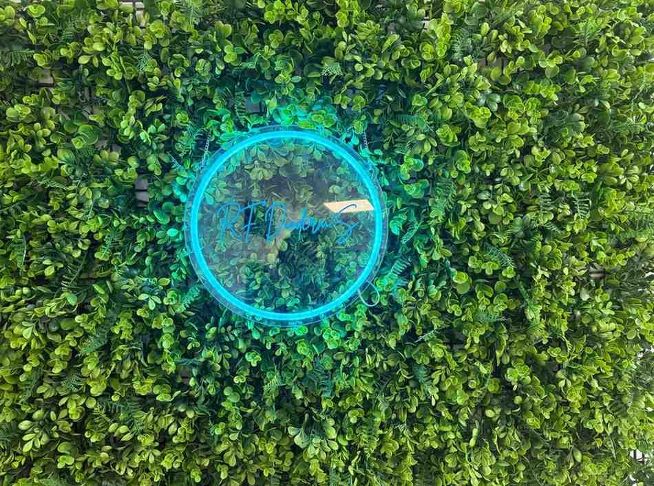 Una pared de follaje artificial de un verde vibrante presenta una luz circular de color azul neón brillante en el centro.