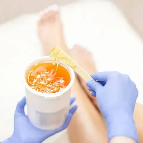 Persona depilándose la pierna. Manos enguantadas sujetando el recipiente y el aplicador de cera. Cera naranja. Cama blanca.