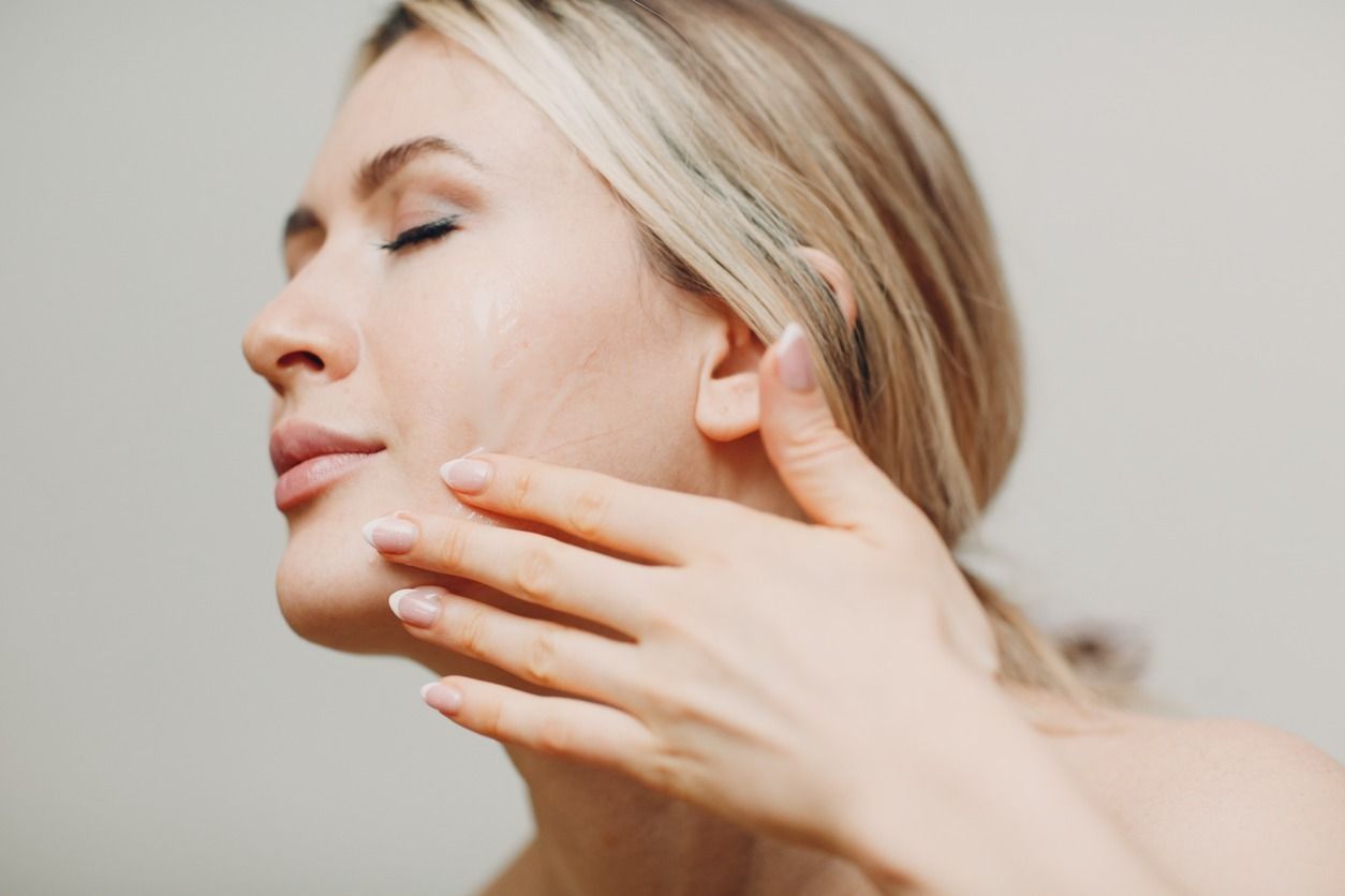 Una persona rubia se aplica suavemente un producto para el cuidado de la piel en la mejilla con una expresión dulce.