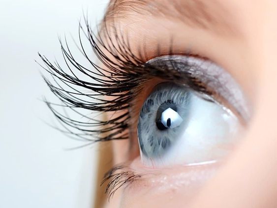 Primer plano de un ojo azul claro con pestañas largas y oscuras, mirando hacia arriba, sobre un fondo pálido.