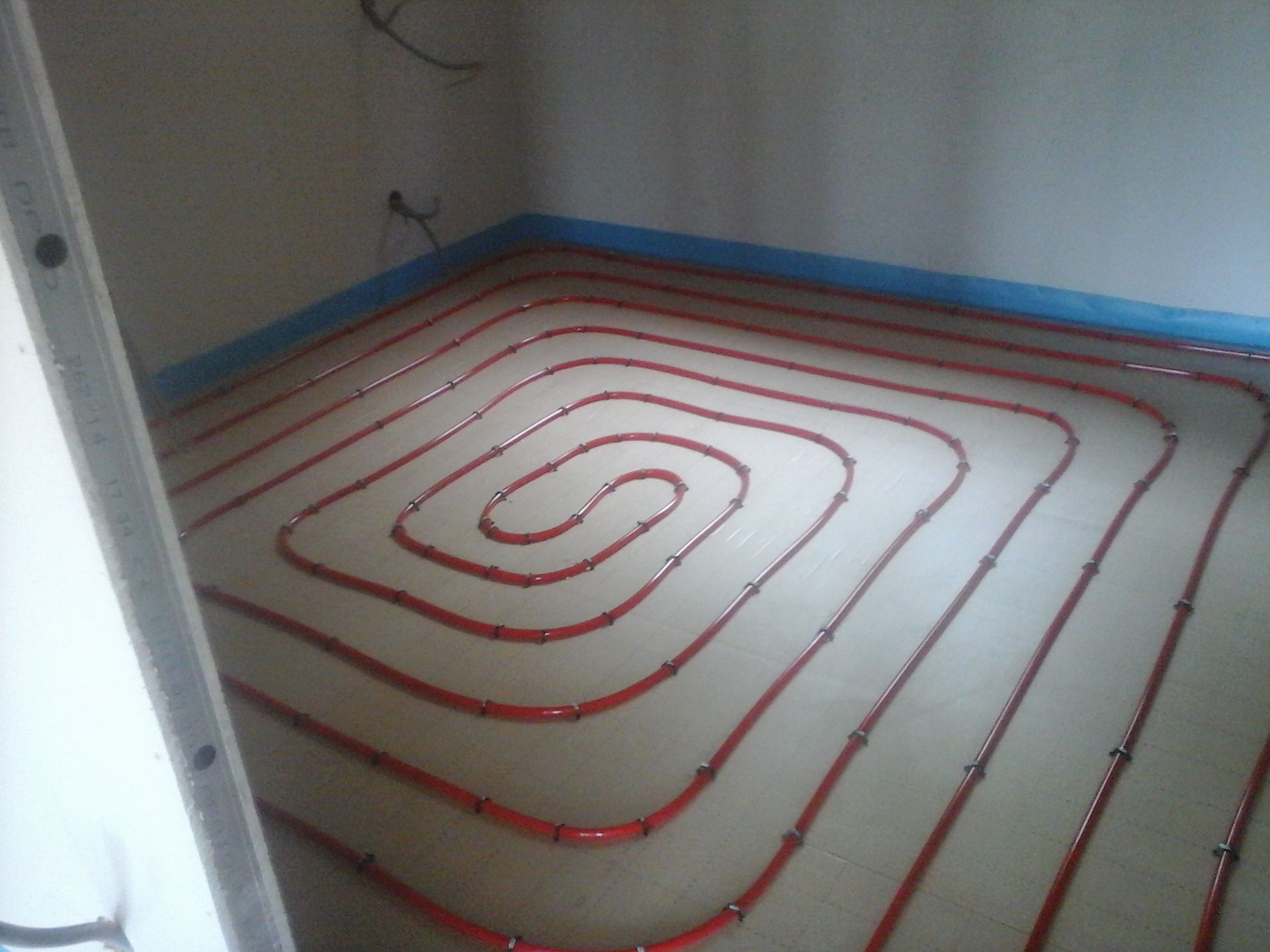 Des tuyaux de chauffage pour sol radiant rouges sont installés en spirale sur un sol bordé de ruban isolant bleu le long des murs.