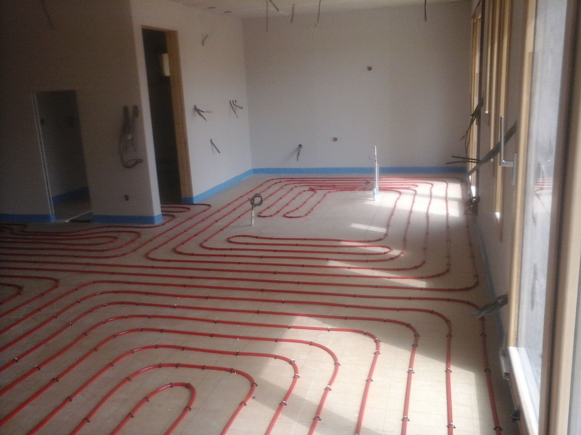 Des tuyaux pour chauffage radiant rouges sont installés en quadrillage sur le sol d'une pièce inachevée aux murs blancs.