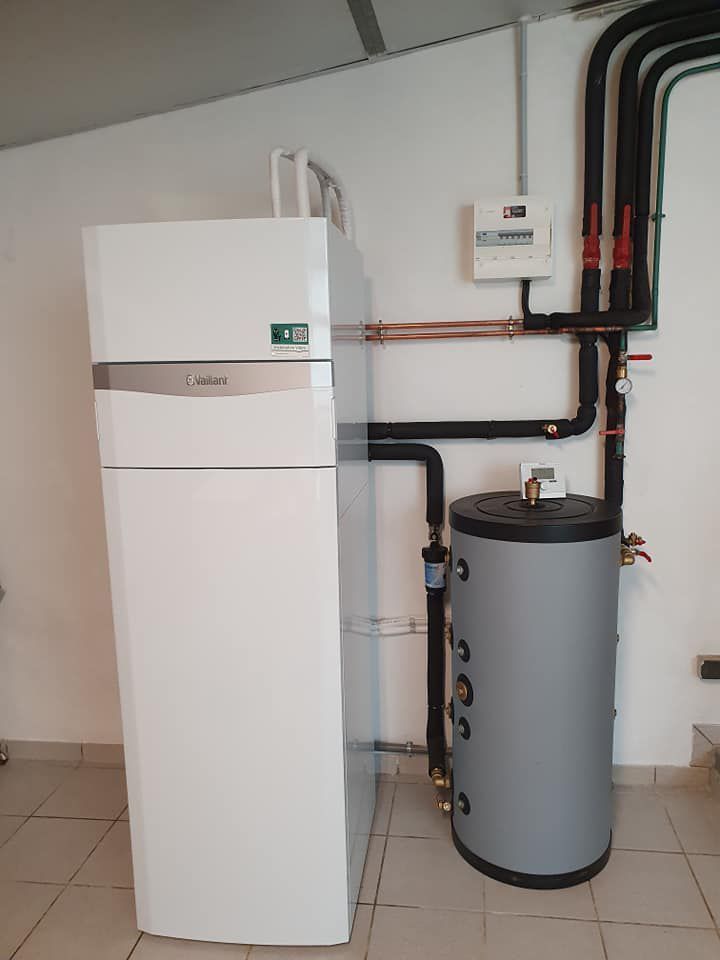 Une pompe à chaleur Vaillant blanche se dresse à côté d'un ballon d'eau chaude cylindrique gris dans une pièce au sol carrelé.