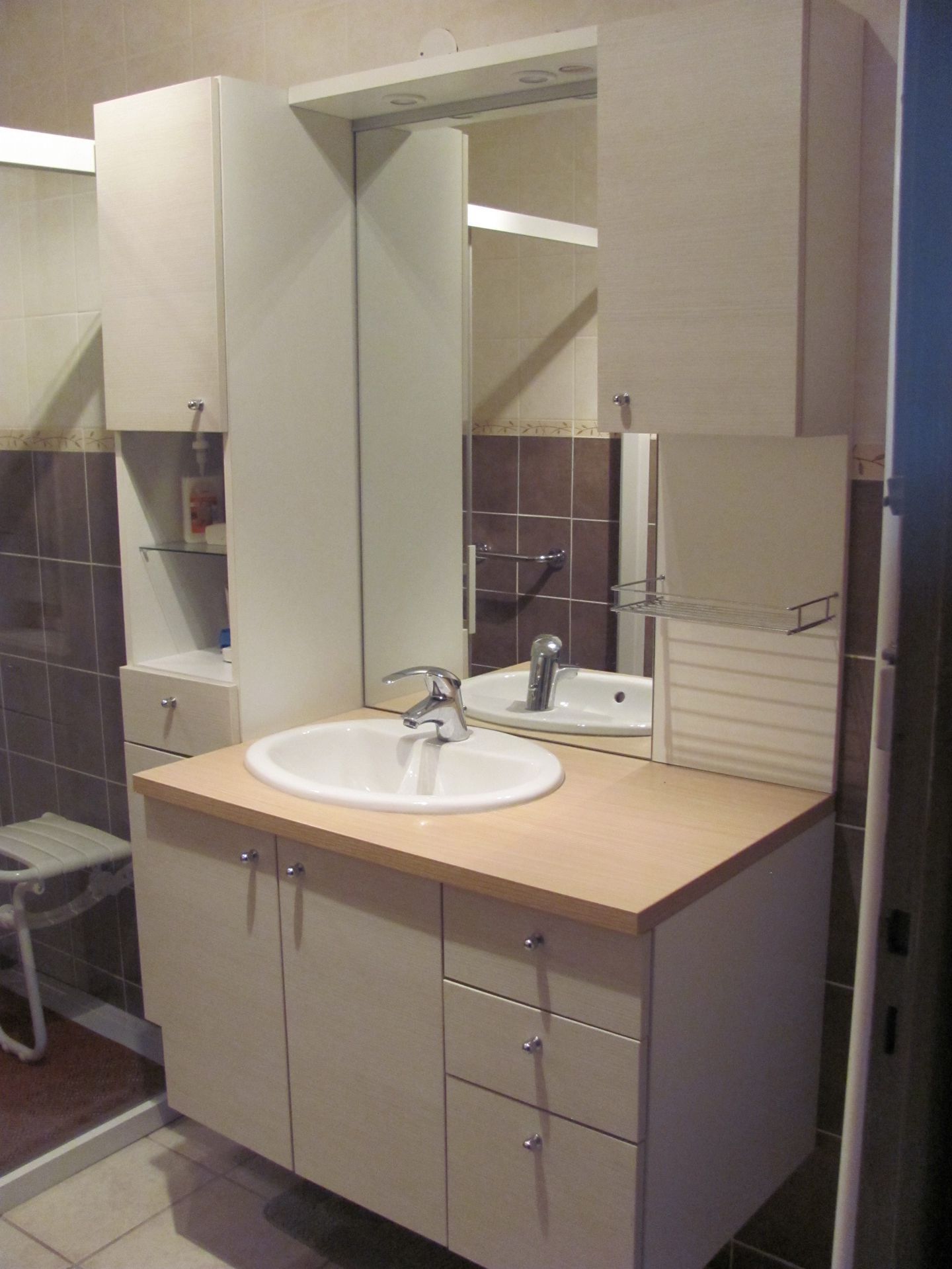 Un meuble lavabo avec un plan de travail en bois clair, des armoires blanches, des tiroirs, un lavabo, un miroir central et des étagères latérales.