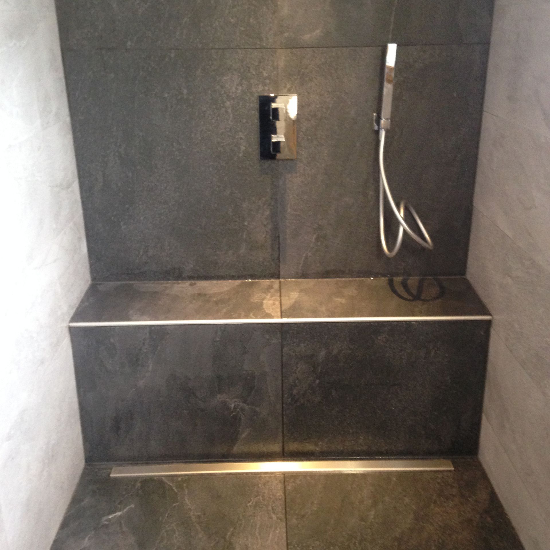 Cette cabine de douche carrelée comprend un banc intégré, une commande de douche murale, une douchette et un caniveau de sol linéaire.