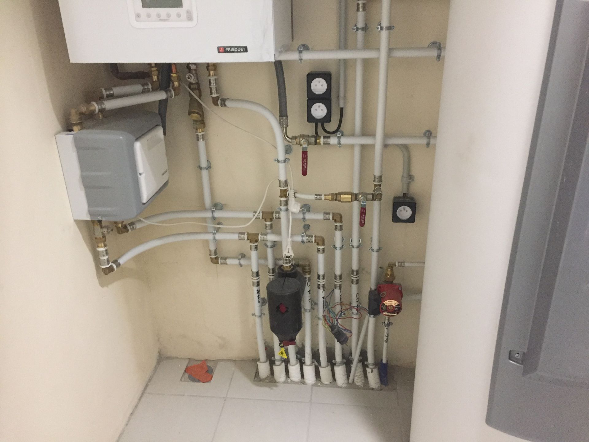 Un placard technique abritant une chaudière murale, des tuyaux blancs, une pompe à eau et diverses vannes de régulation, le tout contre des murs carrelés.