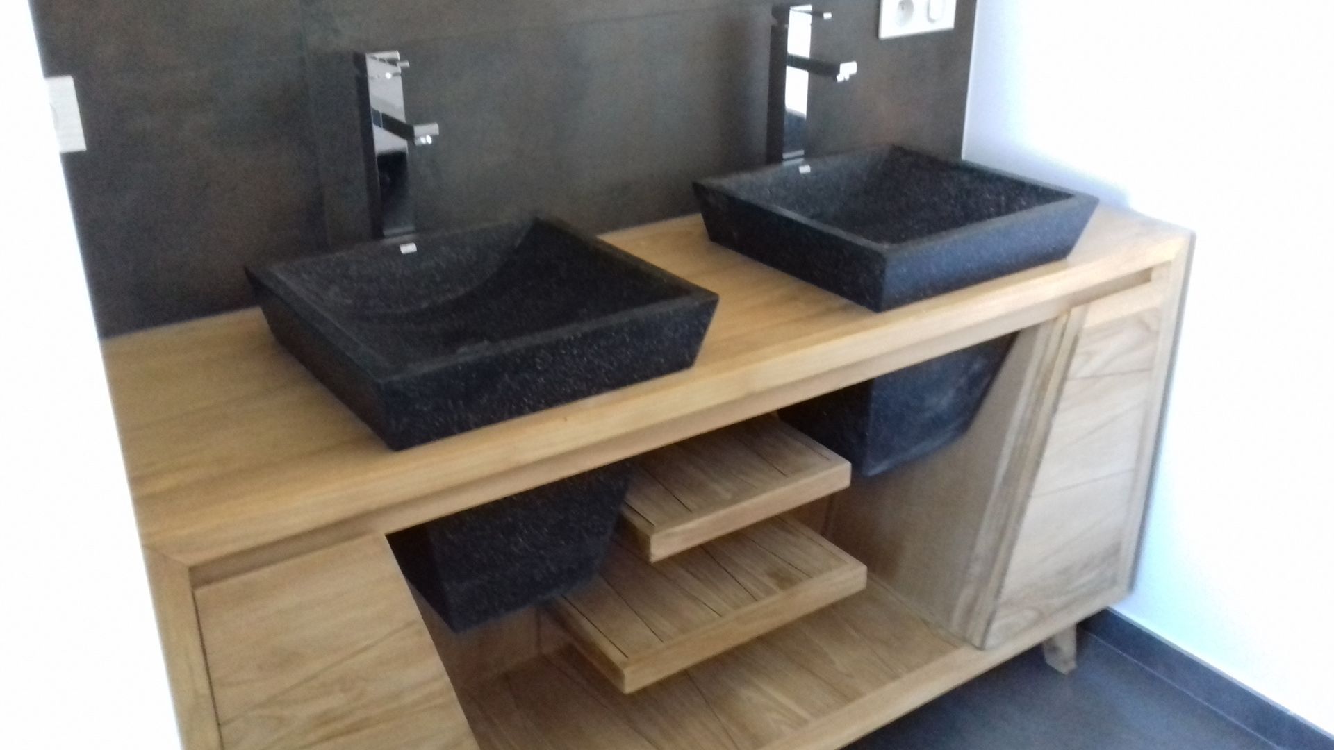 Meuble lavabo moderne avec deux vasques carrées en pierre noire sur un plan de travail en bois avec étagères et placards de rangement.