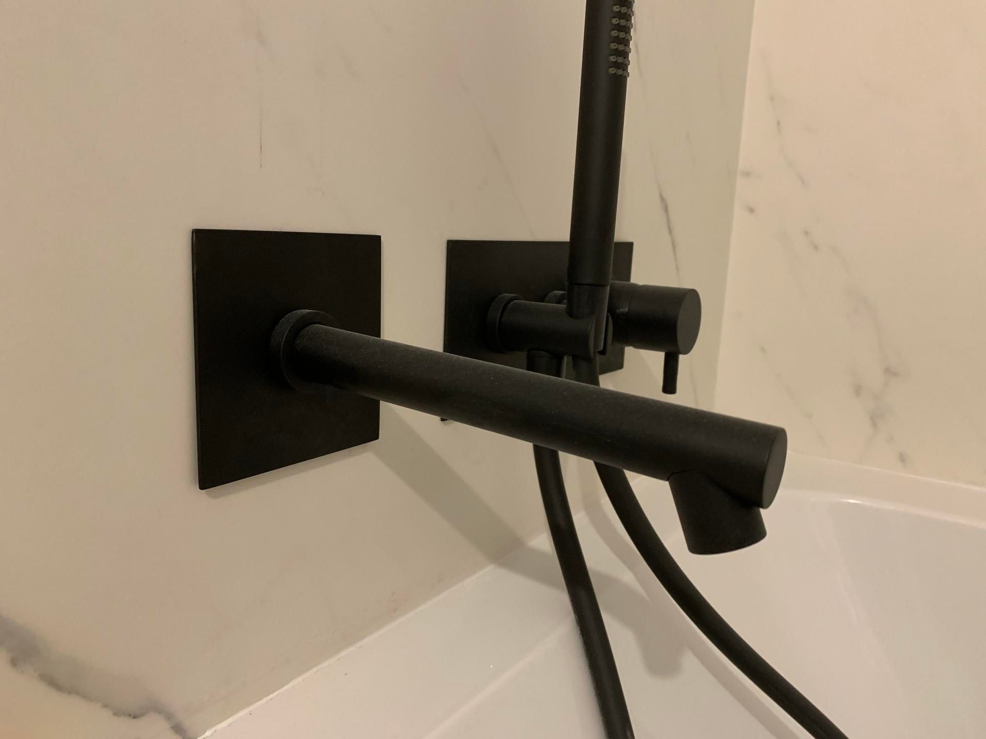 Robinet de baignoire mural noir mat et douchette à main sur un mur carrelé imitation marbre blanc.