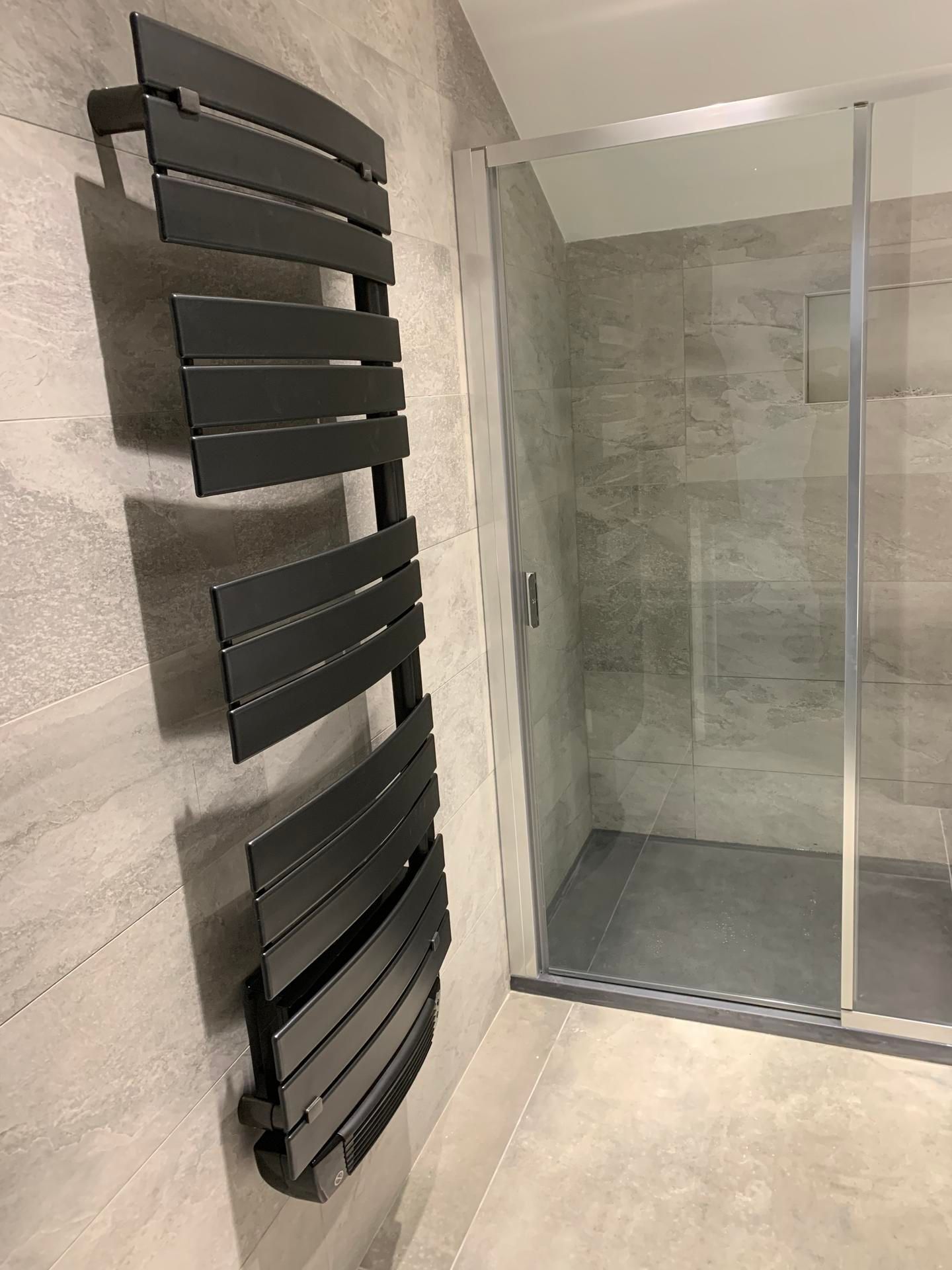 Un sèche-serviettes incurvé moderne, de couleur anthracite, fixé sur un mur carrelé de salle de bains, à côté d'une douche vitrée.