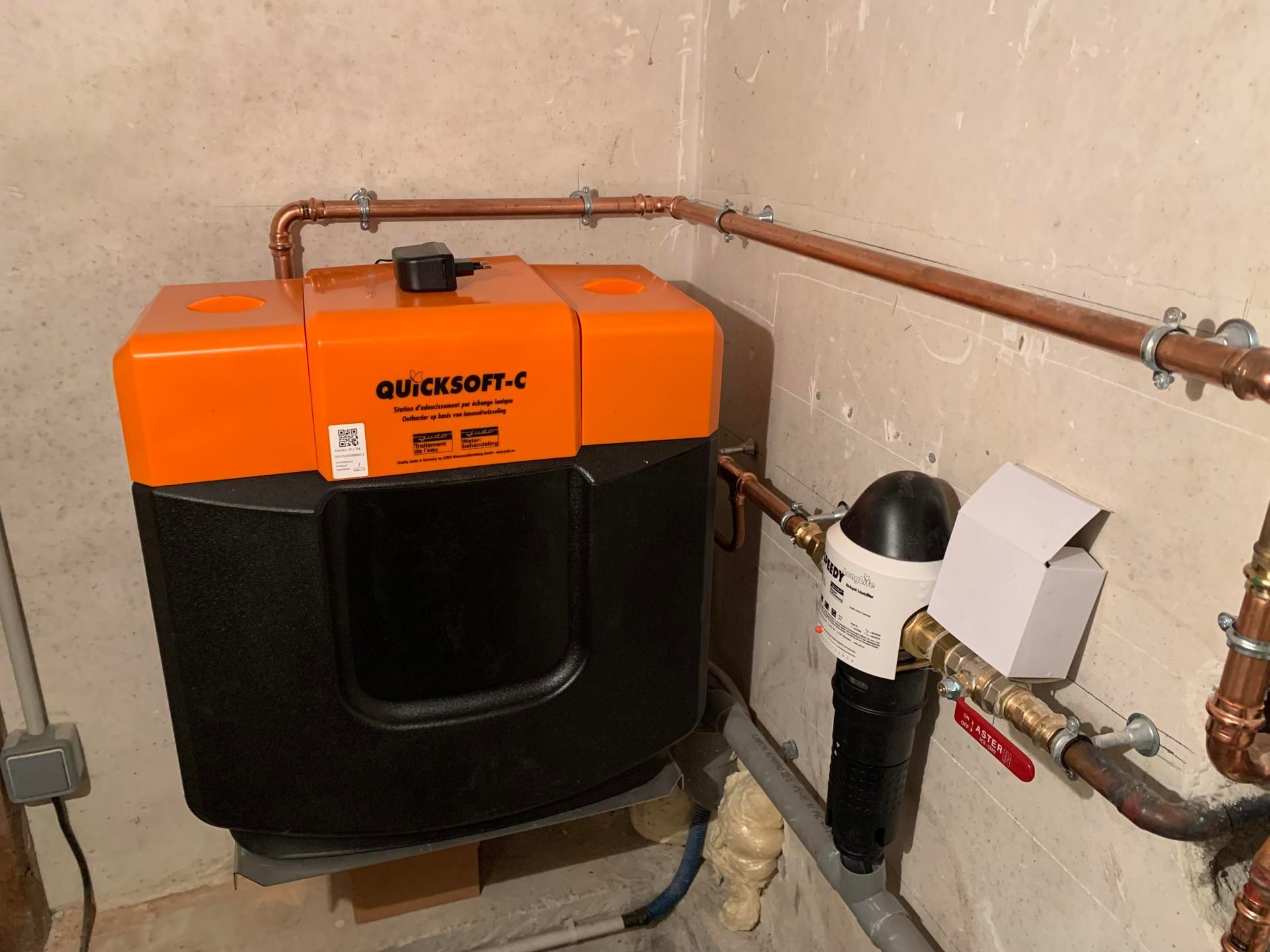 Un adoucisseur d'eau avec un boîtier noir et orange installé dans une pièce avec des tuyaux en cuivre et un filtre.
