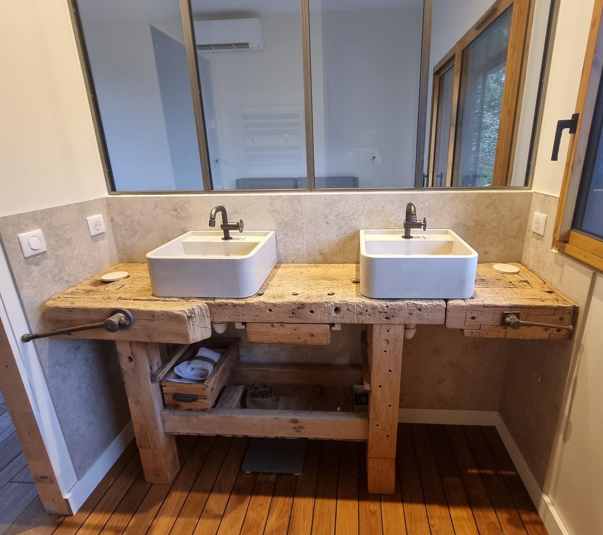 Deux vasques installées sur un établi rustique en bois dans une salle de bains moderne avec un grand miroir et un sol en bois.
