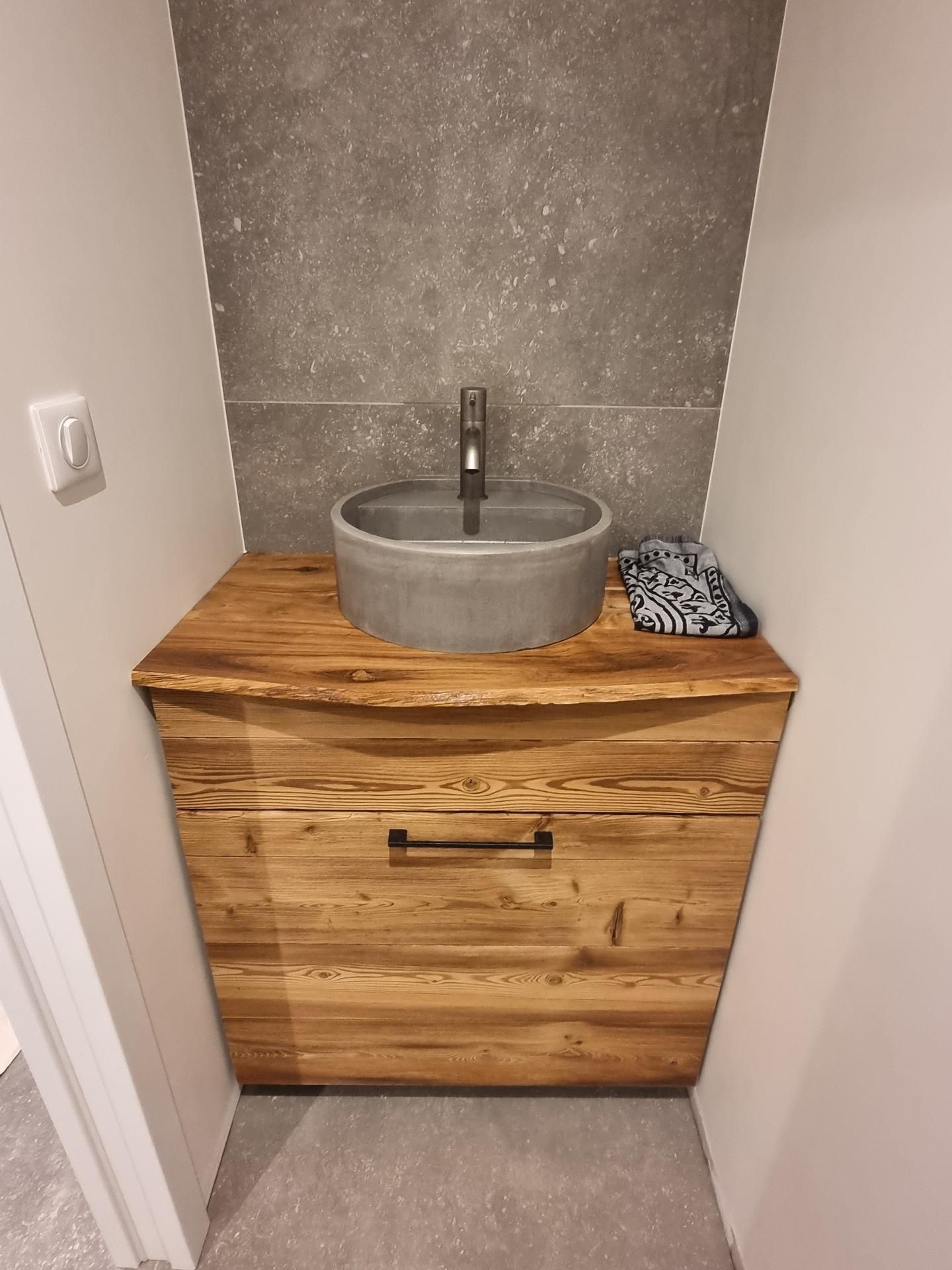 Un meuble lavabo moderne pour salle de bains, comprenant un meuble en bois rustique, une vasque en pierre grise et un robinet foncé sur un carrelage gris.