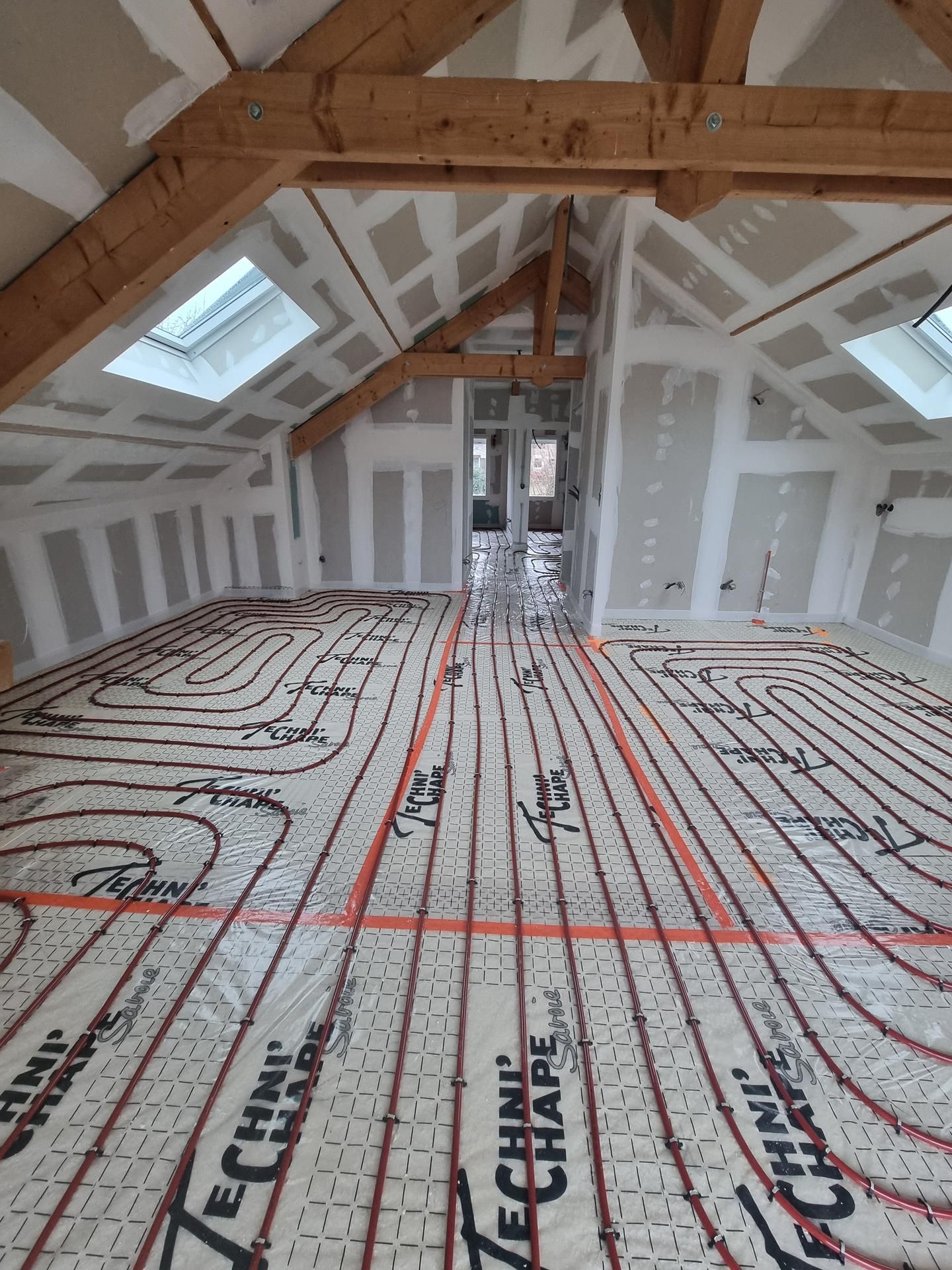 Aménagement d'un grenier avec poutres apparentes, murs en plaques de plâtre et tuyaux de chauffage au sol installés.