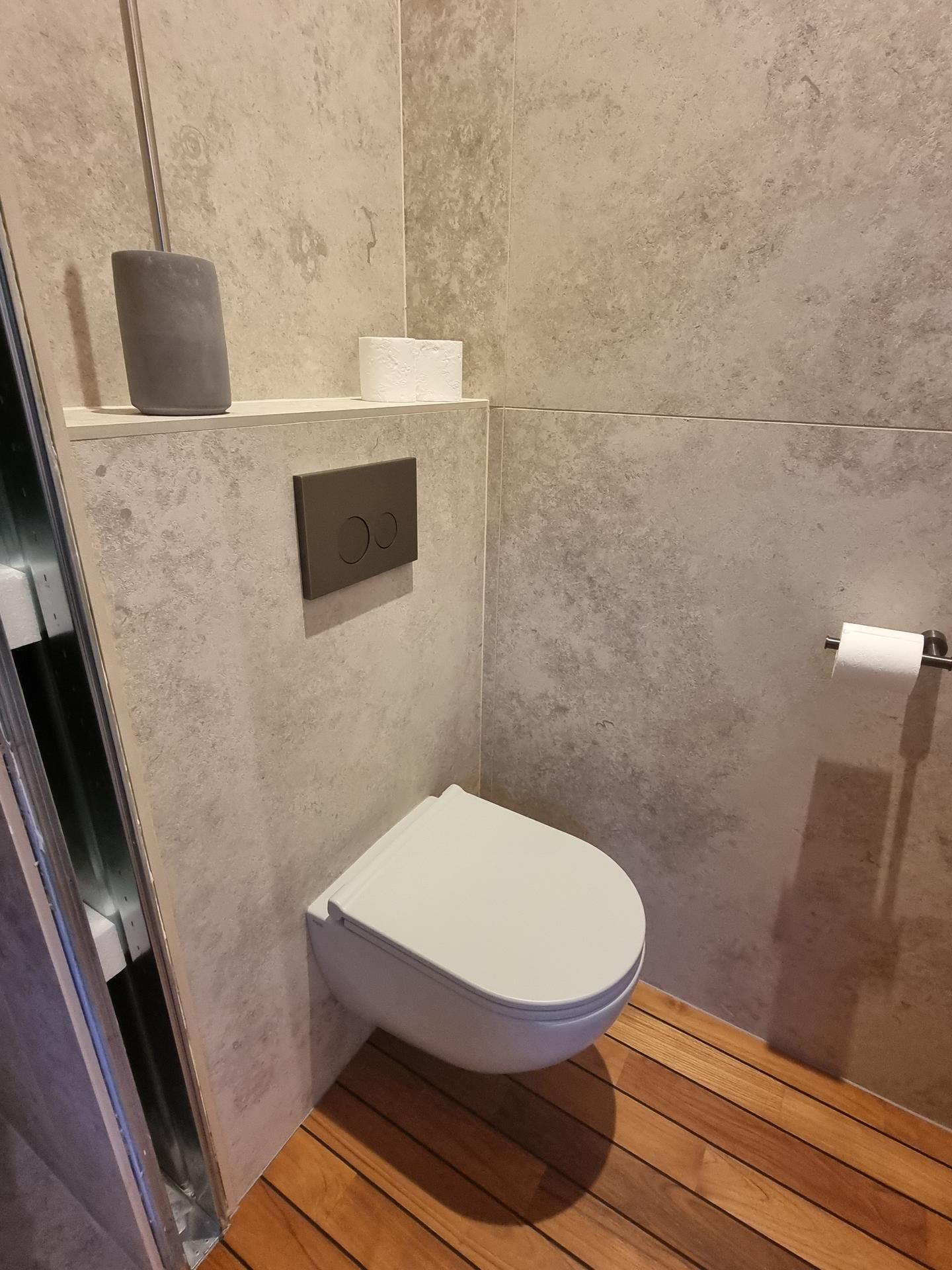 Des toilettes suspendues dans une salle de bains moderne aux murs gris texturés et au sol en lattes de bois.