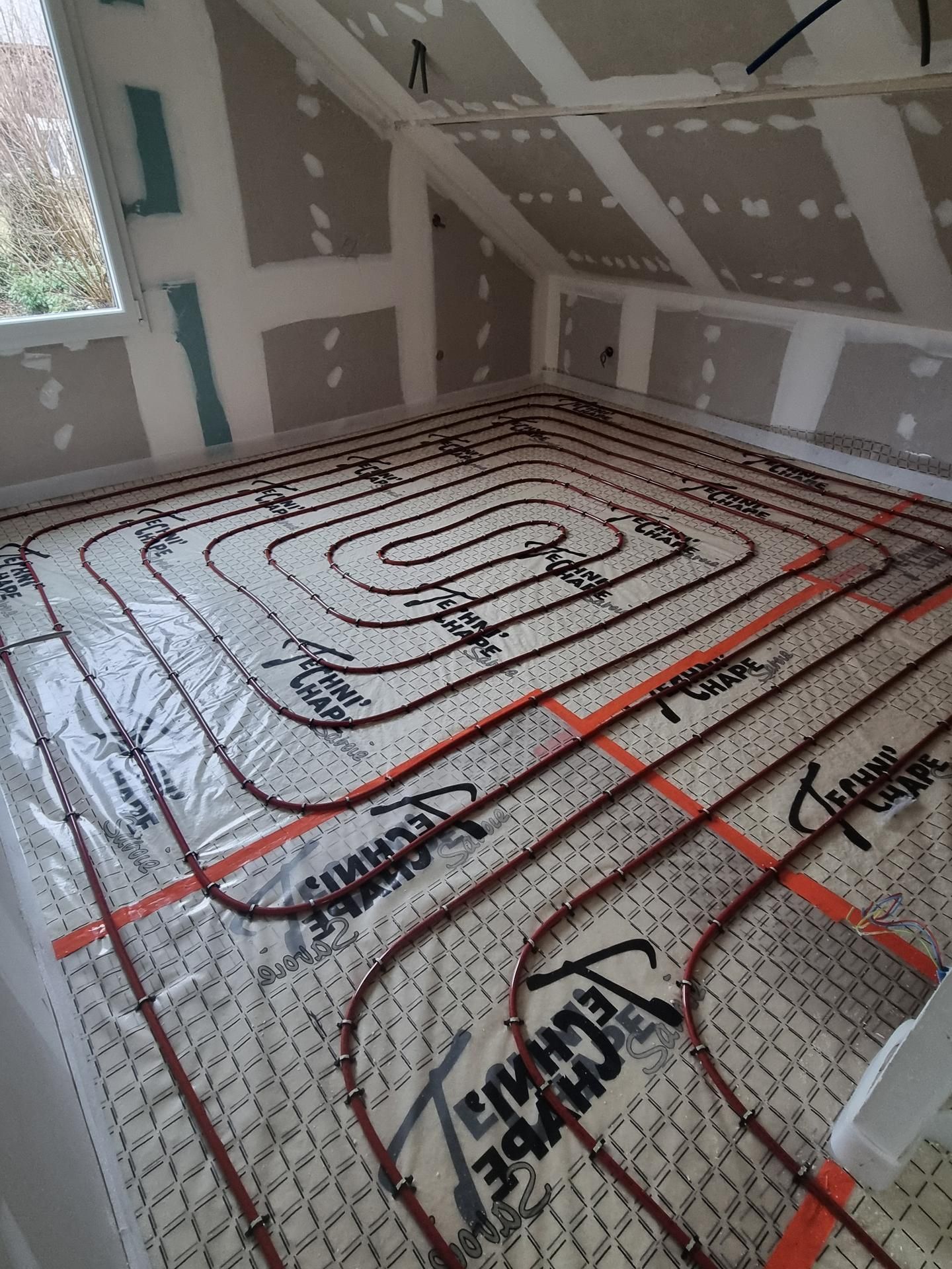 Des tuyaux de chauffage pour sol radiant rouges sont installés en spirale sur un tapis isolant dans une pièce aux murs recouverts de plaques de plâtre.