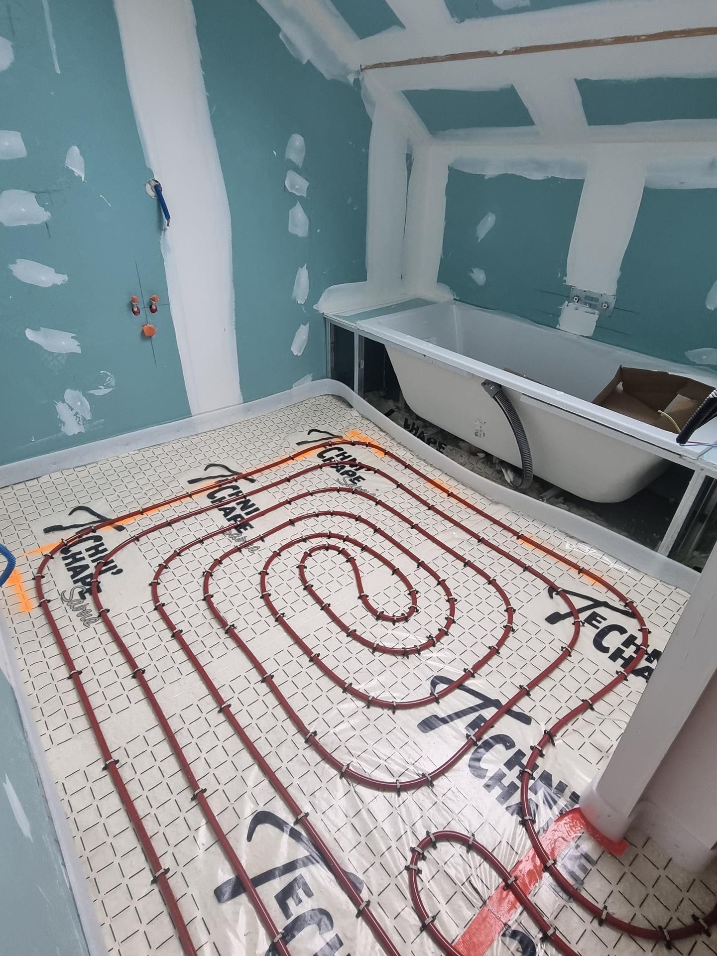 Des serpentins de chauffage au sol rayonnants disposés en grille dans une salle de bains en construction avec une baignoire blanche.