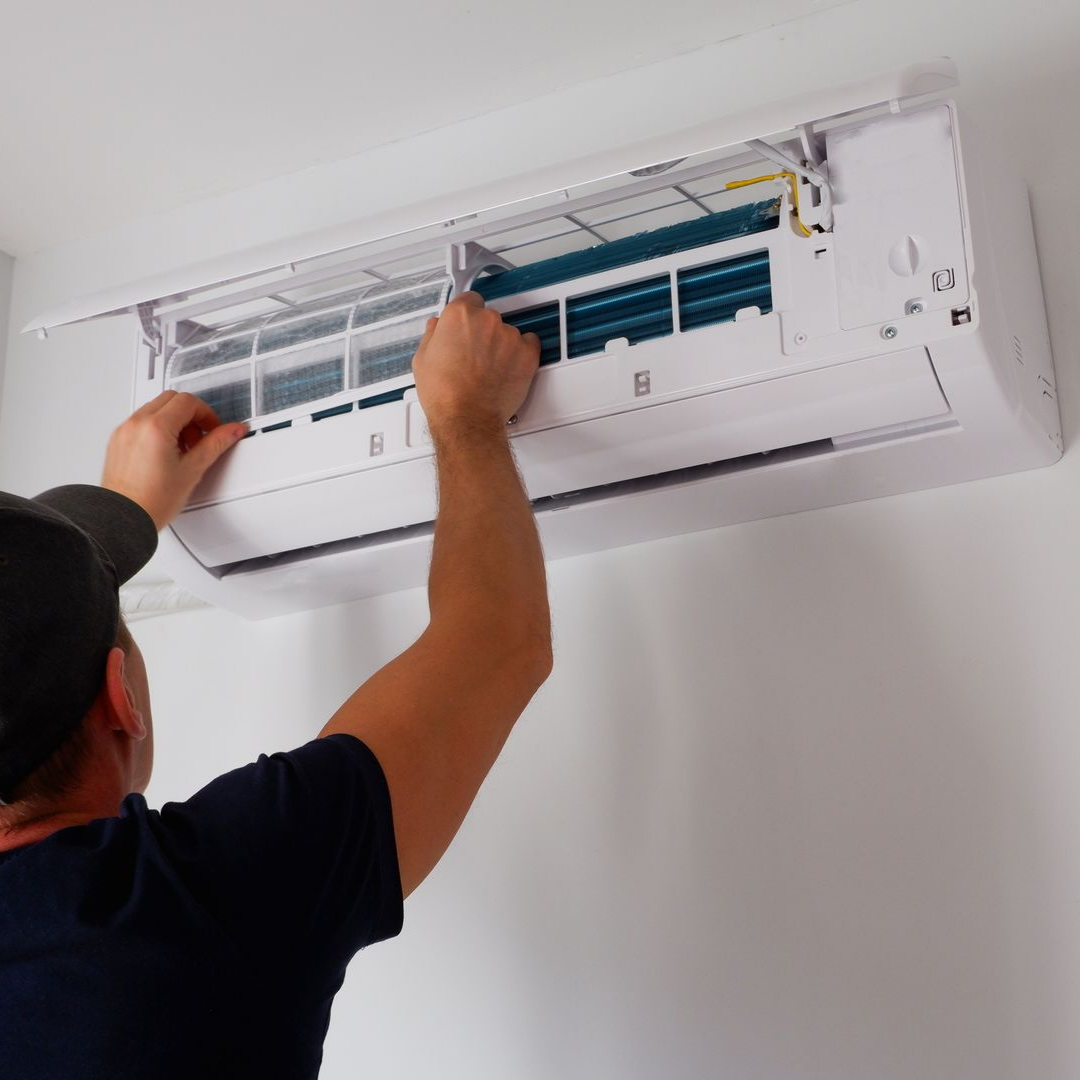 Une personne portant une chemise sombre et une casquette tend la main pour retirer le filtre d'un climatiseur mural.