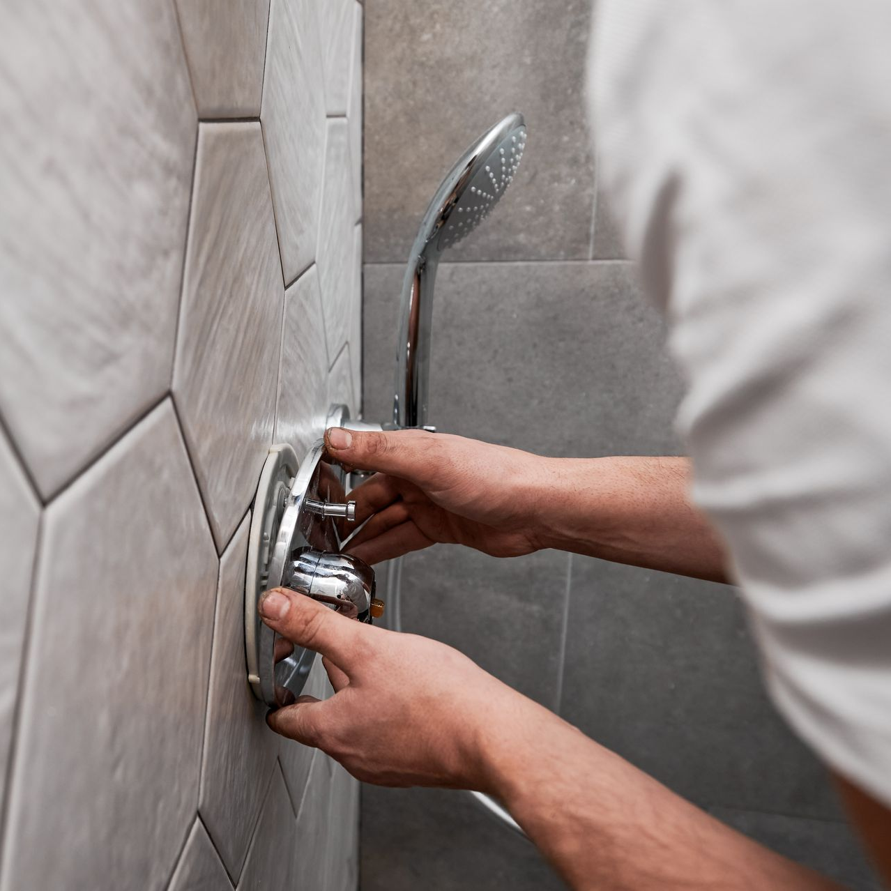 Un plombier installe un robinet de douche et une douchette contre un mur carrelé recouvert d'hexagones de couleur claire.
