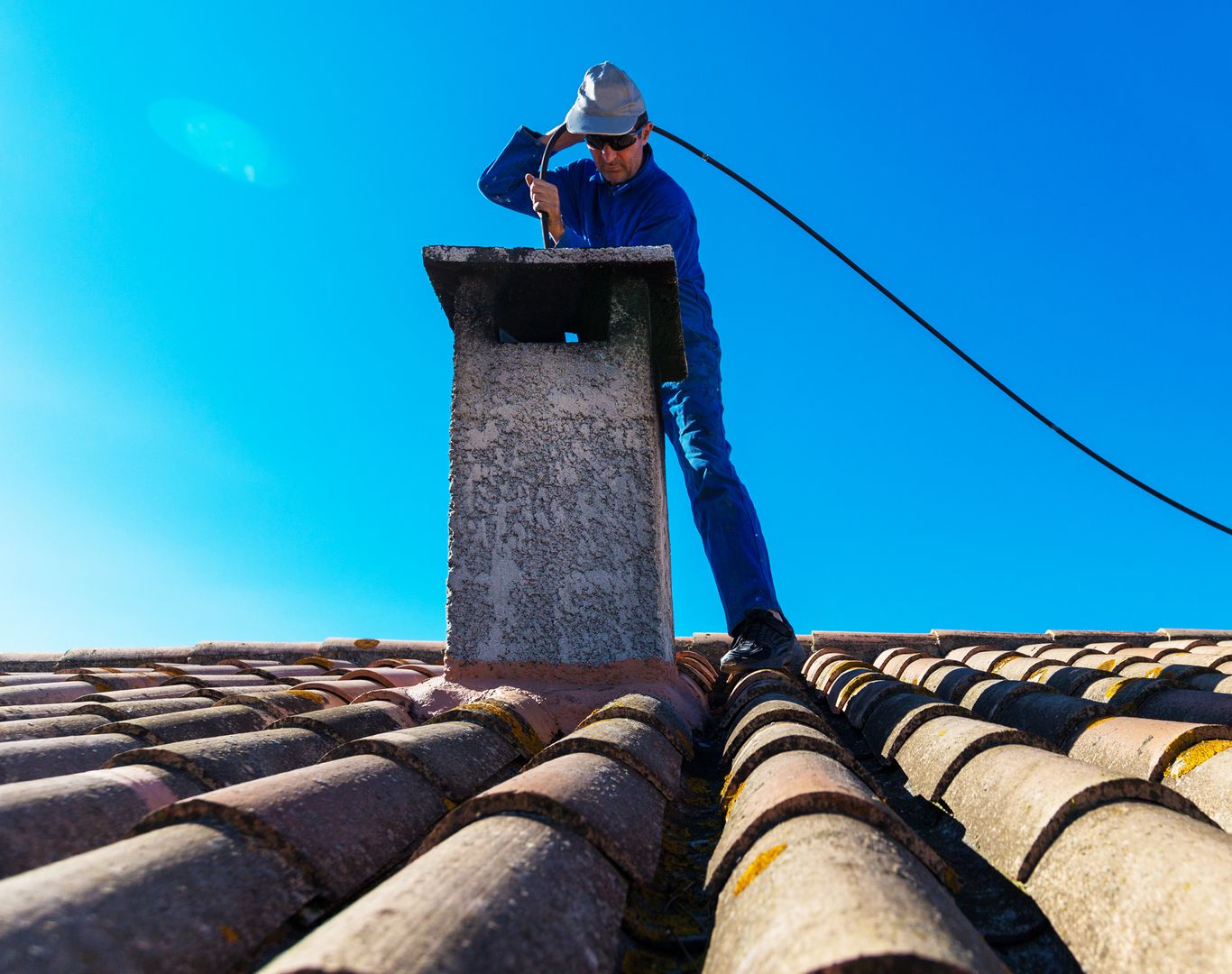Une personne en combinaison bleue nettoie une cheminée sur un toit de tuiles sous un ciel bleu dégagé.