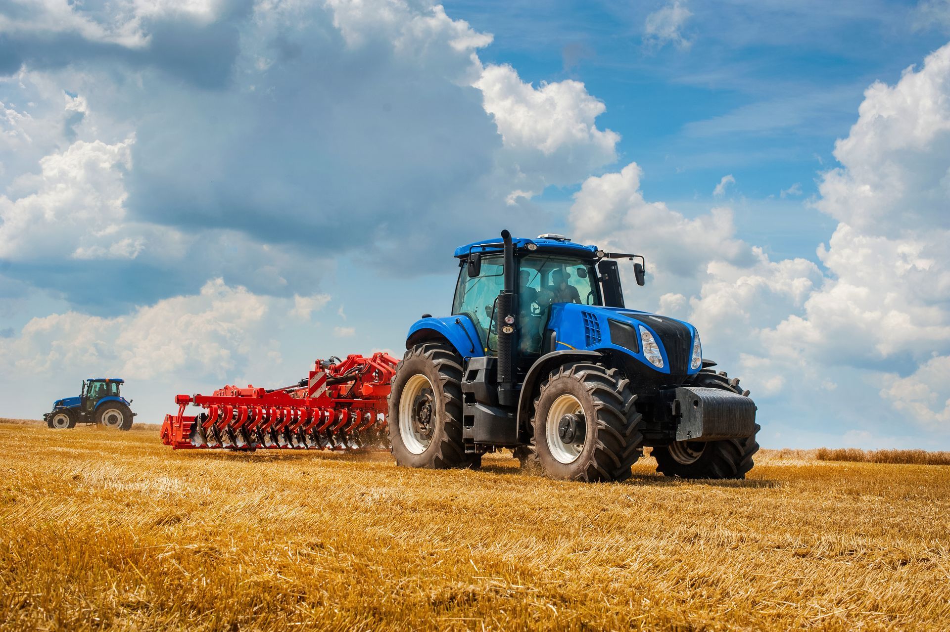 Un tracteur bleu tirant un semoir rouge à travers un champ doré moissonné, sous un ciel bleu parsemé de nuages ​​blancs duveteux.