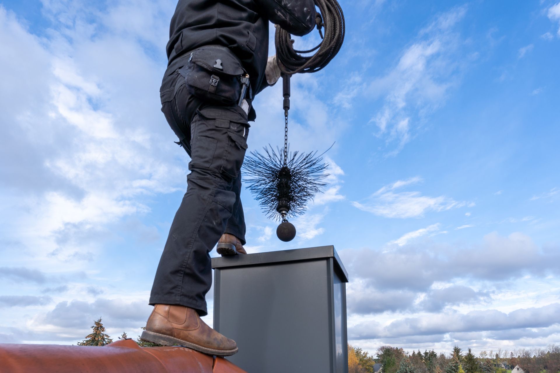 Un ramoneur, sur un toit, descend une brosse ronde dans une cheminée, sur fond de ciel bleu nuageux.