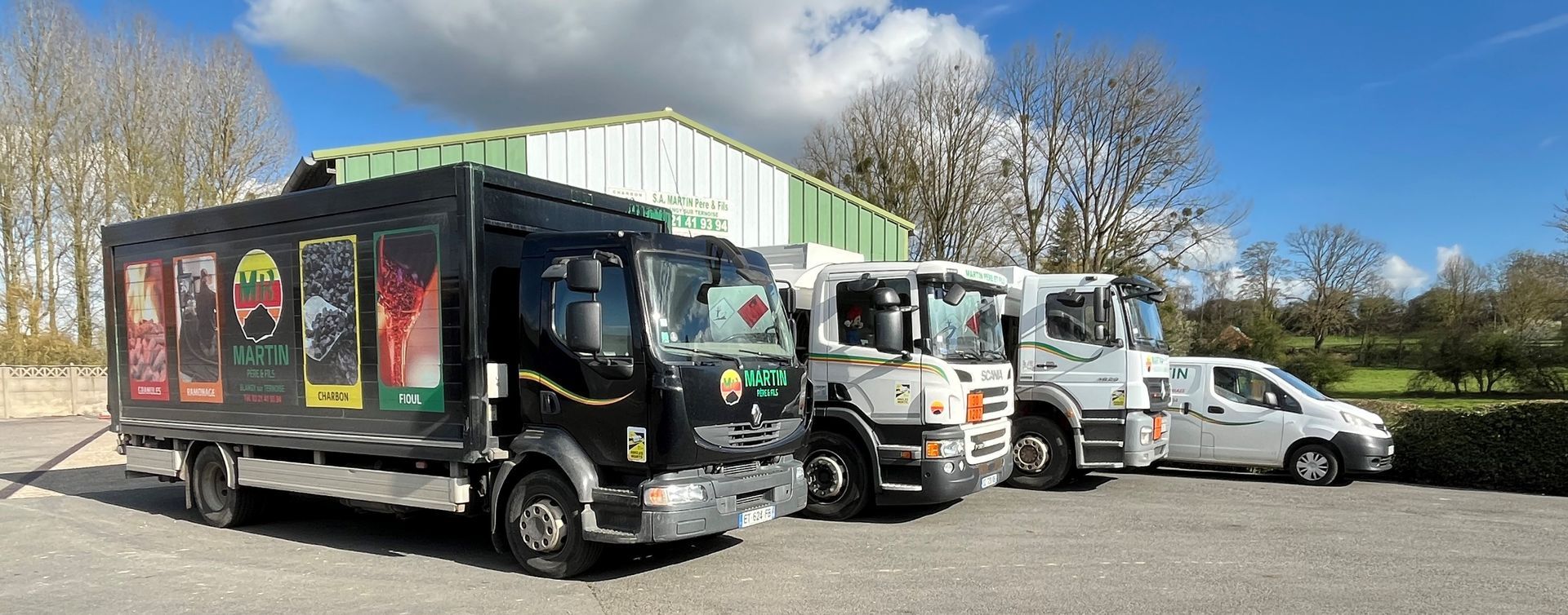 Une rangée de camions garés et une camionnette blanche stationnée sur un terrain gravillonné devant un entrepôt, sous un ciel bleu.