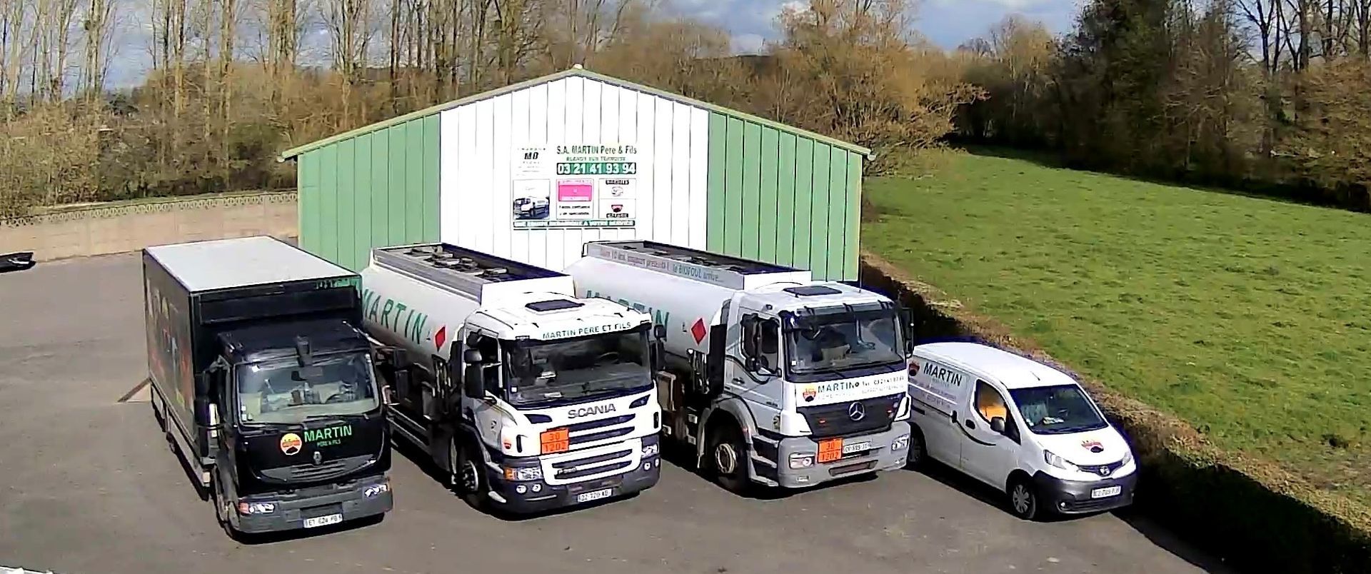 Quatre véhicules de l'entreprise, dont deux camions-citernes, un camion noir et une fourgonnette blanche, étaient stationnés devant un hangar vert.