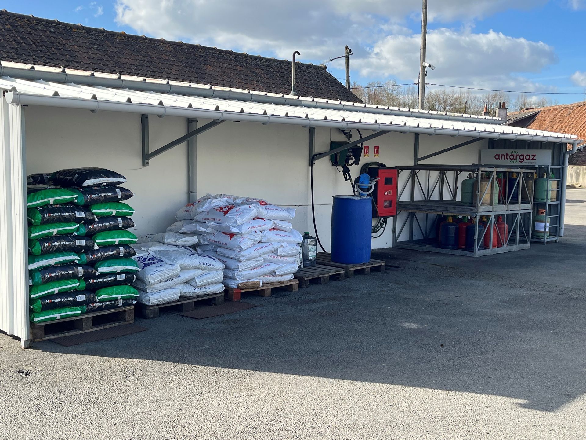 Un hangar de stockage extérieur contient des palettes de produits ensachés, un fût en plastique bleu et un support métallique avec des bouteilles de gaz.