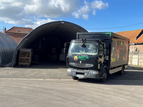 Un camion de livraison noir est garé sur un terrain gravillonné devant un grand hangar de stockage métallique en forme d'arche, sous un ciel bleu ensoleillé.