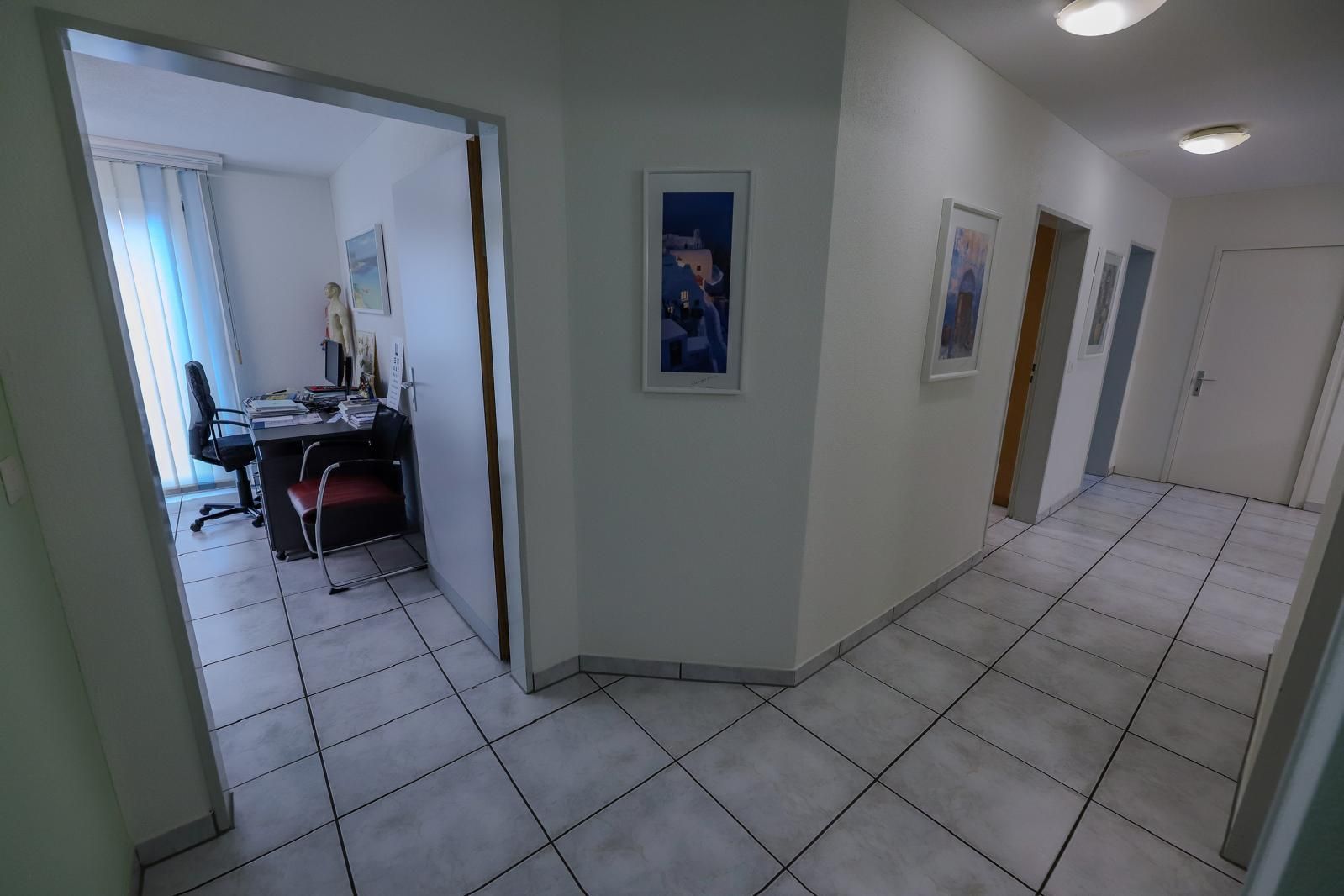 Flur mit weißem Fliesenboden, offener Bürotür links, gerahmten Bildern an den Wänden und geschlossenen Türen geradeaus