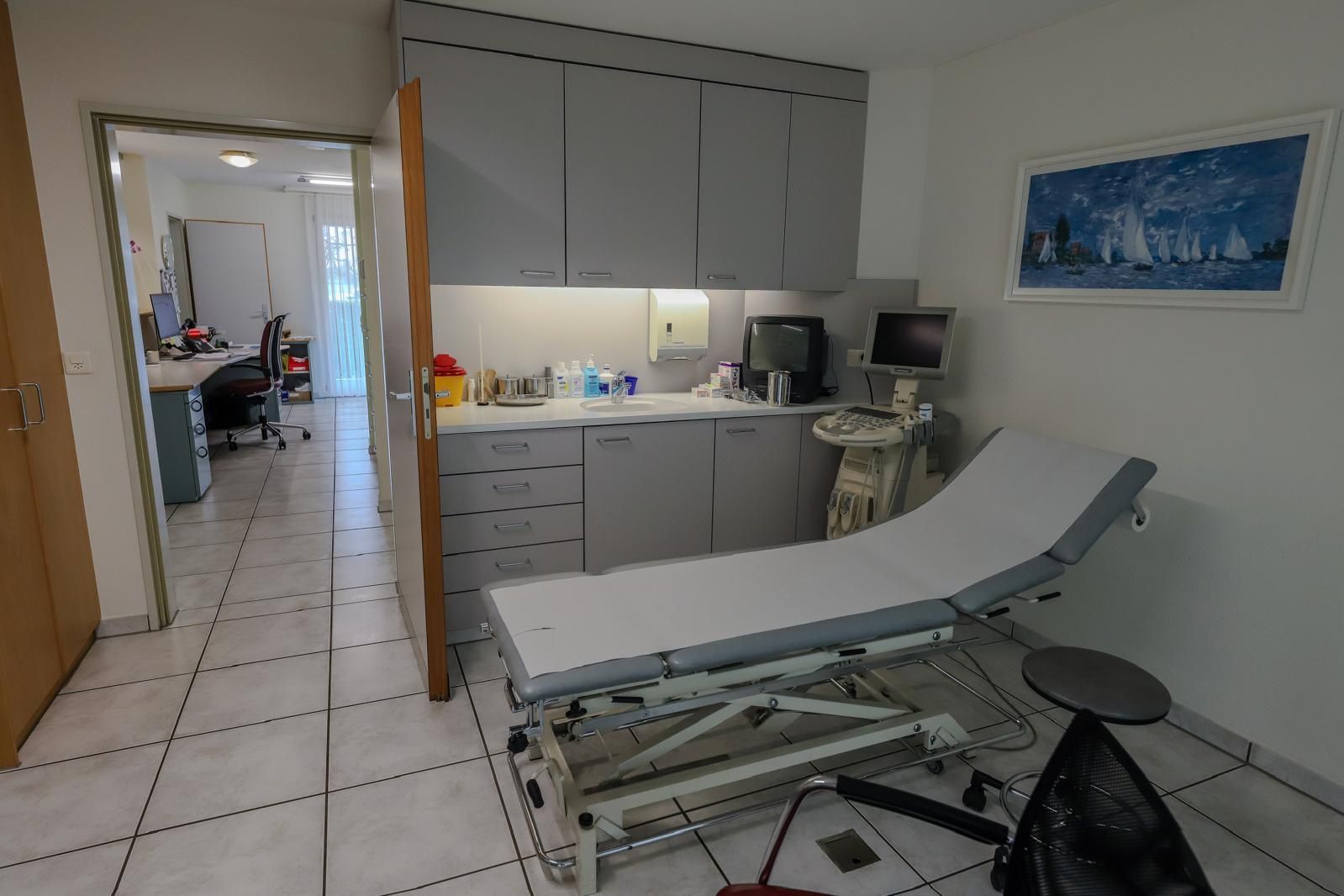 Untersuchungsraum mit Behandlungsliege, Schränken, Waschbecken und Fliesenboden in einem Praxisbüro