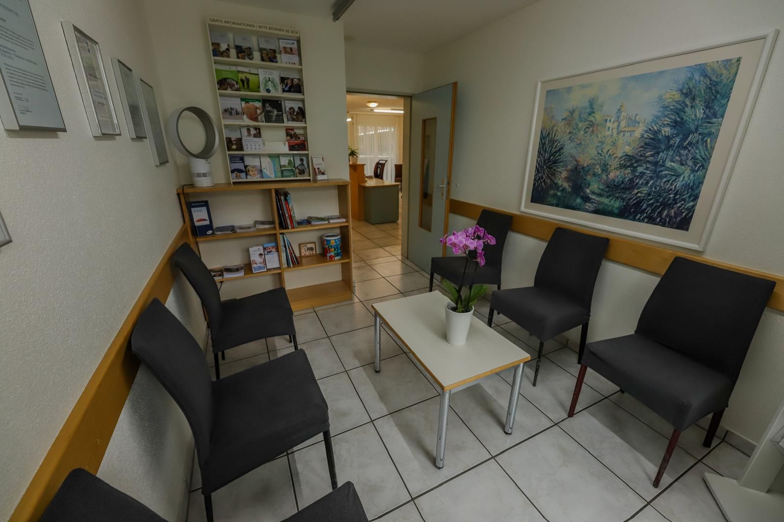Kleiner Warteraum mit schwarzen Stühlen, weißem Tisch, Fliesenboden, Regal und floraler Wandkunst