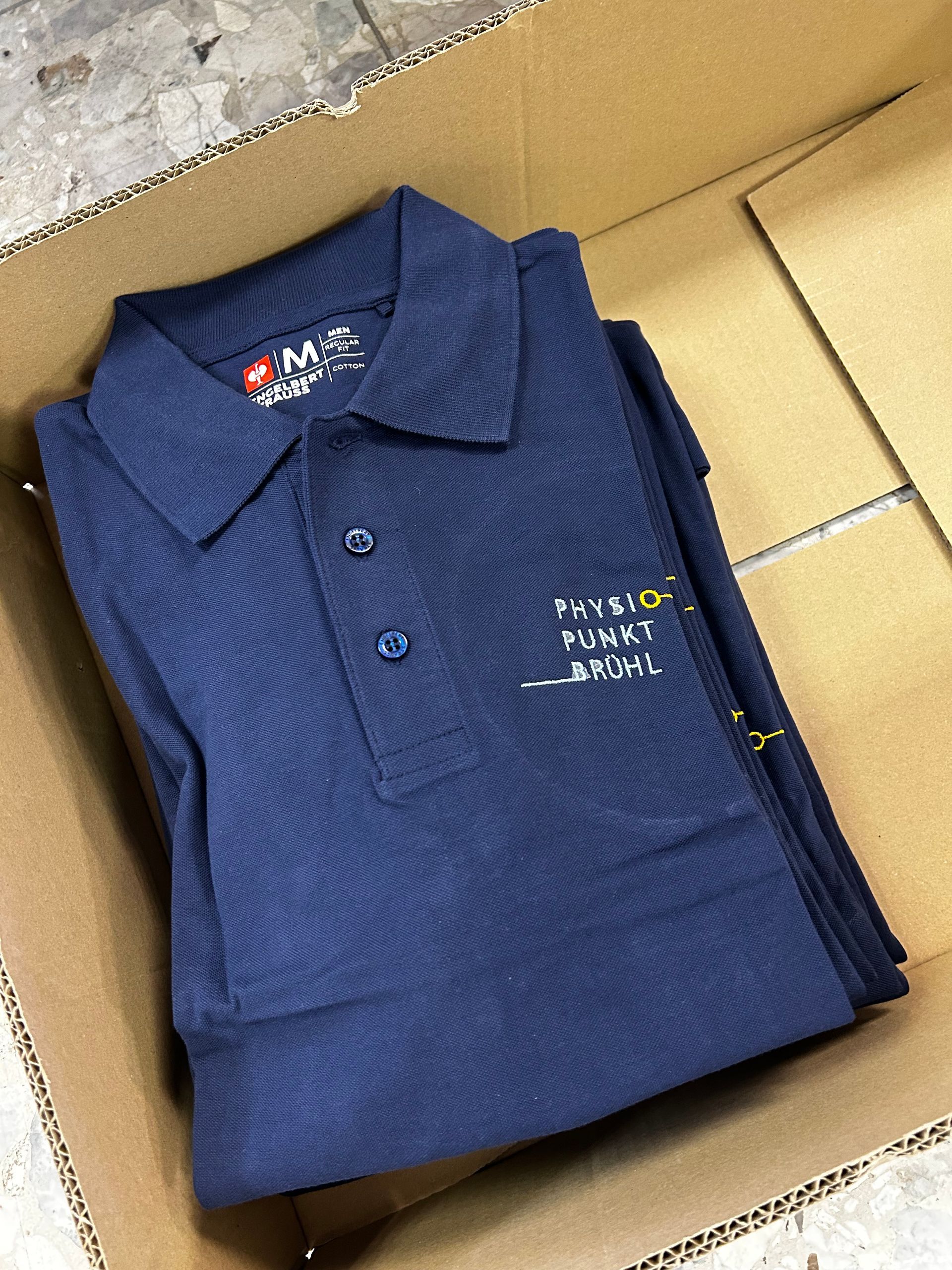 Folded navy-blue polo shirt in a cardboard box, with white embroidered text on the chest.