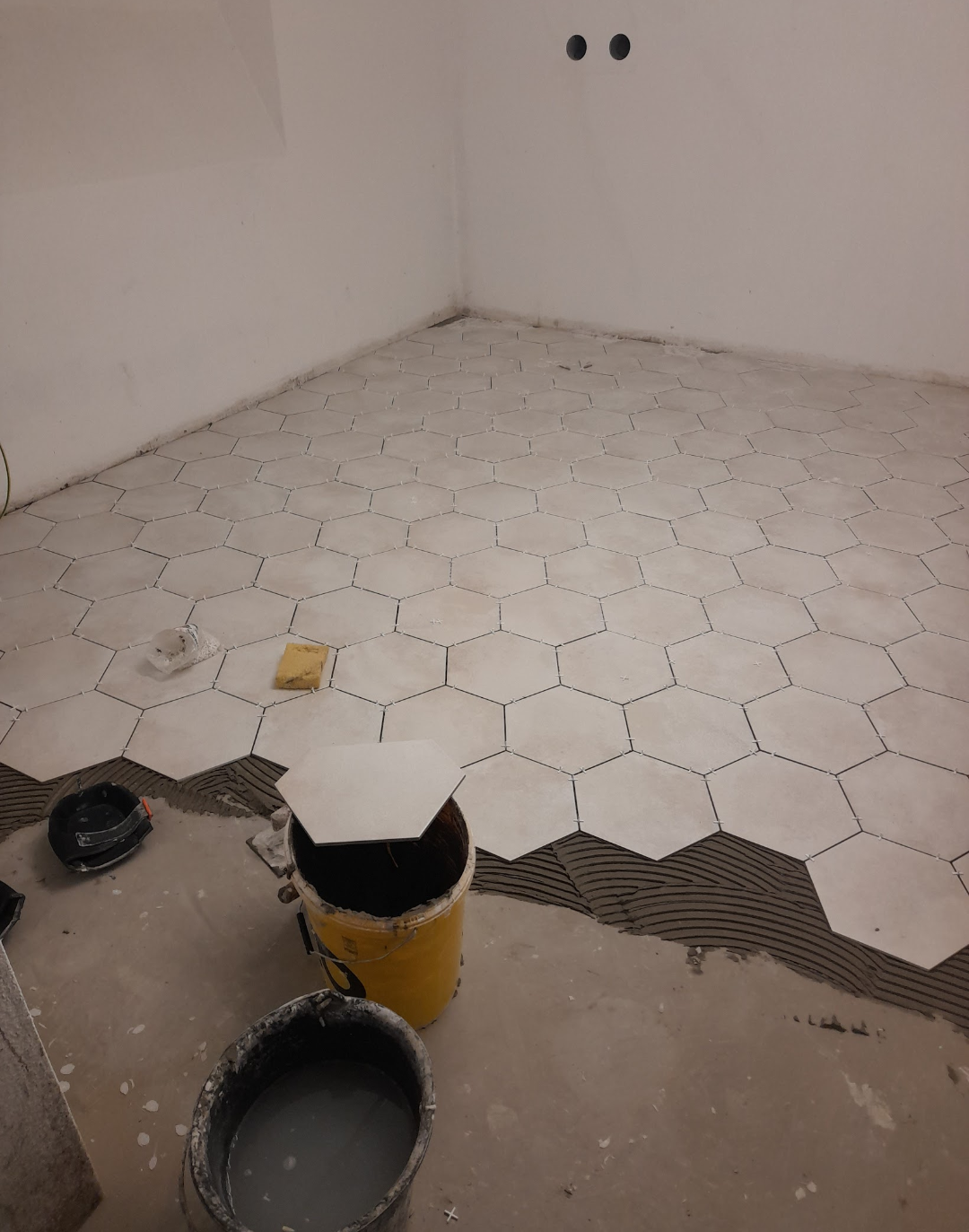 Des carreaux hexagonaux sont posés sur un sol en béton dans une pièce en construction, avec des seaux et des outils visibles.