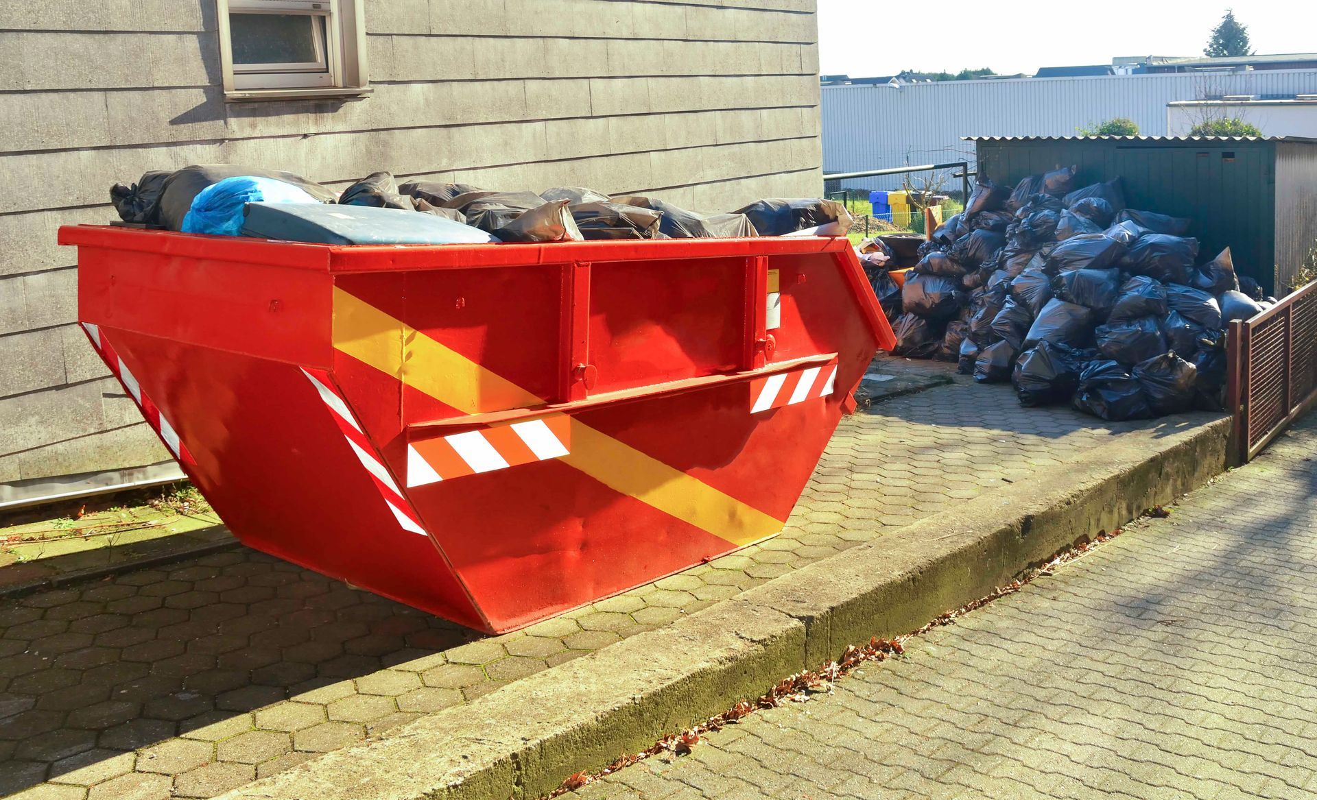 Ein roter Müllcontainer steht neben einem Stapel Müllsäcke.