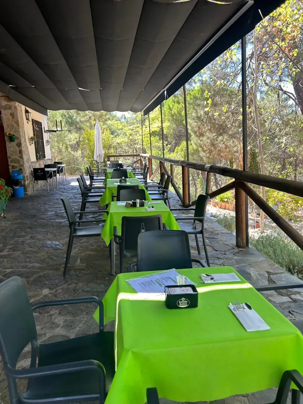 Restaurante al aire libre con manteles verdes, sillas negras y una barandilla de madera con vistas a los árboles.