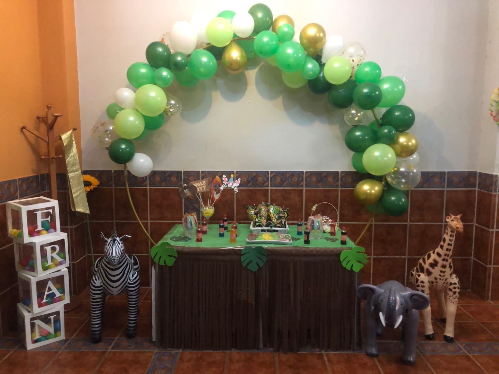 Decoración de fiesta con temática de safari con arco de globos, pastel, cebra, elefante, jirafa y letras mayúsculas.