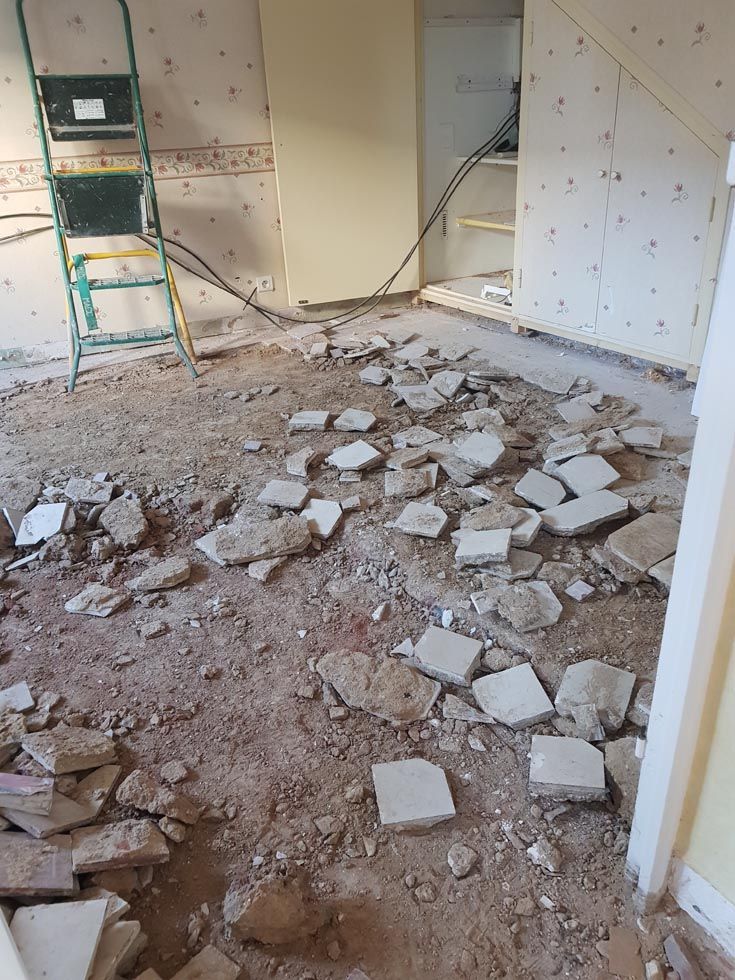 Pièce en rénovation, morceaux de carrelage éparpillés sur le sol poussiéreux, échelle appuyée contre le mur.