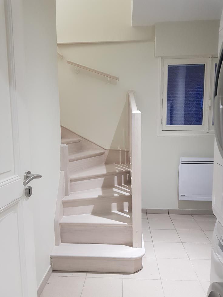 Escalier avec marches et rampe claires. Une fenêtre et un lave-linge se trouvent à droite.