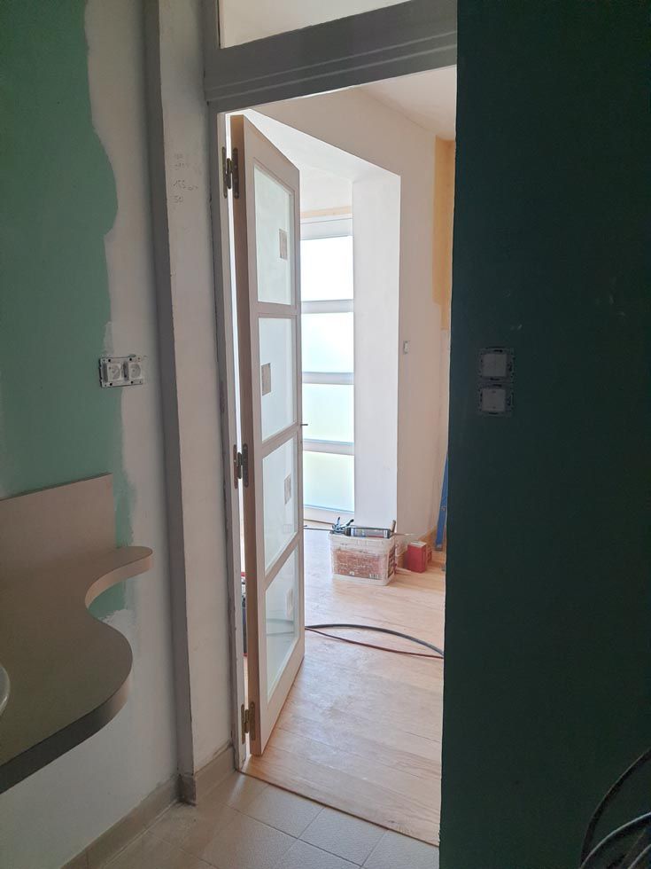 Porte ouverte donnant sur une pièce avec fenêtre. Porte en bois clair à panneaux de verre. Murs verts et blancs.